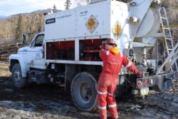 A worker in red coveralls and a hard hat is operating a drilling rig mounted on a white truck in an outdoor setting with trees in the background.