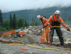 Two construction workers in orange high-visibility clothing are managing cables on a muddy construction site with an excavator operating in the background amidst a forested area with low clouds or mist.
