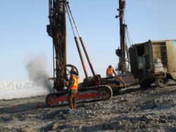 Workers in high-visibility vests operating heavy drilling machinery on a site with snow in the background.