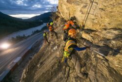 Workers conduct rock stabilization above a road during twilight, secured with ropes and harnesses for safety.