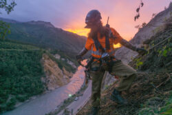 A worker in safety gear uses a rope for support while standing on a steep slope, with a river below and a sunset in the background.