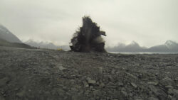 A controlled explosion taking place in a rugged, mountainous landscape, possibly for construction or mining purposes, with a plume of smoke and debris rising from the ground.