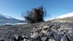 An explosion generates a cloud of debris along a rocky lakeshore with snow-capped mountains in the background.