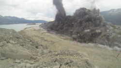Explosion in a quarry with debris and dust flying through the air, with a body of water and a ship in the background.