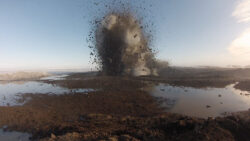 A geyser eruption in a muddy field with debris being thrown into the air against a clear sky.