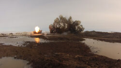 Explosion occurring in a muddy field with debris and a fireball visible.
