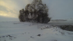 A large explosion ejecting smoke and debris creates a cloud above the snowy ground, with the blast appearing to have originated from a point on the surface.