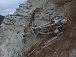 Construction workers operate a drilling rig on a steep rock face, secured with safety harnesses and ropes.