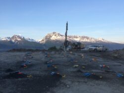 Geotechnical drilling rig at a mountainous field site with survey flags and equipment scattered around.
