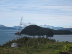 A lattice transmission tower stands on a hill overlooking a scenic view of a coastline with forested islands and mountainous terrain in the background.