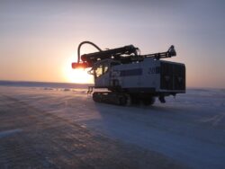 A tracked snow vehicle equipped with a large drill at sunset on a vast icy landscape.