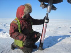A person in cold weather gear is kneeling on snow, inspecting a piece of equipment, possibly for ice drilling or sampling.
