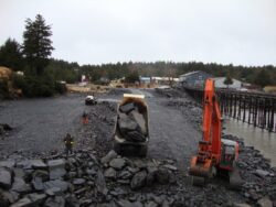 A large excavator is loading a haul truck with rocks at a construction site near a body of water with a wooden pier and buildings in the background.