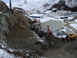 Construction machinery operating near a rock excavation site with buildings in the background.
