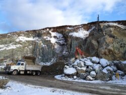 A dump truck and excavator at a mining or quarry site with large chunks of excavated material on the ground, under a clear sky with patches of snow visible on the ground.
