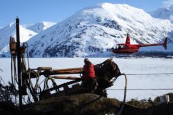 A worker operates drilling machinery on snowy terrain with a red helicopter flying in the background against a backdrop of snow-covered mountains.