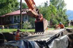 Construction workers in high visibility vests are using heavy equipment to excavate and perform work on an outdoor construction site near a residential building.